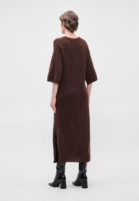 Brūns adījuma kleita ar brīvu piegriezumu, īsām piedurknēm un sānu šķēršļiem. Kombinēta ar melniem augstiem zābakiem uz vienkārša fona.