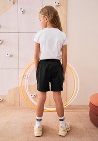 Un enfant portant un t-shirt blanc et un short noir se tient de dos. Caractéristiques notables : ceinture élastique, cerceaux blancs et oranges en arrière-plan.