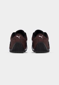 Ζευγάρι καφέ και μαύρα παπούτσια Puma, εμφανιζόμενα από πίσω, με λαστιχένιες σόλες και λευκό λογότυπο Puma σε κάθε τακούνι.