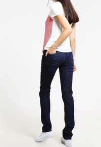 Maglietta bianca con righe rosse su un lato, abbinata a jeans blu scuro, caratterizzati da una vestibilità slim e un classico design a cinque tasche. Scarpe sportive bianche.