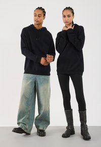 Svart hettegenser i mykt stoff på to personer. Den mannlige personen har på seg løse, lyse denimjeans og svarte støvler; den kvinnelige personen har på seg tights og høye svarte støvler.