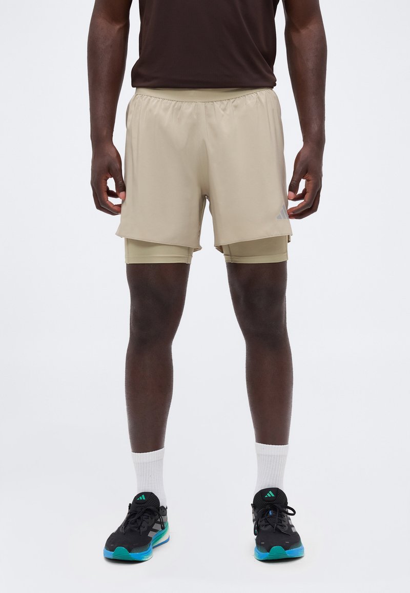 Homme portant un short de sport beige à couches, des chaussettes foncées et des chaussures de course noires avec des semelles bleues et vertes.