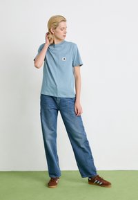 Personne se tenant sur un sol vert, portant un t-shirt bleu, un jean bleu ample et des baskets marron, touchant ses cheveux et regardant sur le côté.