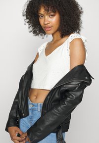 Top cropped branco com rendilhado e alças com babados, usado sob uma jaqueta de motociclista de couro preta, combinado com jeans de cintura alta azul clara.