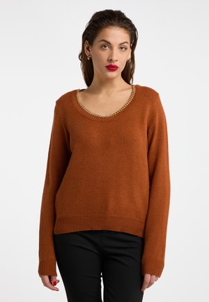 Vrouw met donker haar draagt een burnt orange trui met een gouden kettingdetail bij de hals en zwarte broek tegen een effen witte achtergrond.