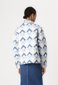 Chaqueta acolchada en un patrón geométrico azul y blanco, con un cuello puntiagudo y puños con botones, combinada con una falda de mezclilla.