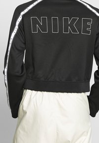 Černá mikina s dlouhým rukávem Nike se bílými pruhy "AIR" na rukávech a velkým konturovaným textem "NIKE" na horní části zad, nošena se světlemi kalhotami.