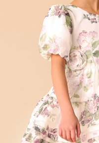 Cvetlična obleka s puffnimi rokavi, narejena iz lahkega blaga. Vzorec vključuje svetlo roza in zelene cvetlične motive ter dekorativni cvetlični dodatek.