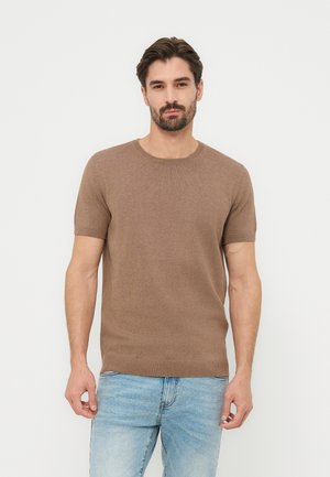 Muškarac u smeđem džemperu kratkih rukava i svijetloplavim trapericama stoji okrenut prema naprijed na jednostavnoj svijetloj pozadini.