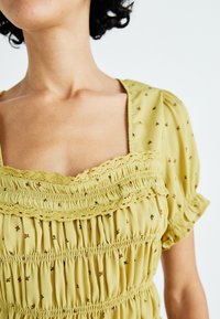 Geel gesmokt blouse met korte pofmouwen, voorzien van kantafwerking en kleine bloemenpatronen door het hele shirt. Lichtgewicht stof met een gestructureerde afwerking.