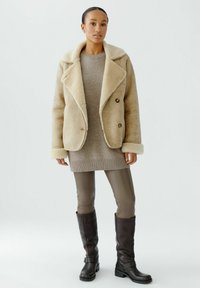 Beige schapenvachtjas met een brede kraag, gedragen met een lichtbruine trui, strakke taupe leggings en kniehoge donkerbruine leren laarzen.