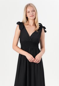 Vestido preto com um decote em V profundo, cintura franzida e alças de tecido presas em laços nos ombros. Textura suave e silhueta fluida.