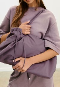 Žena drži veliki mekani ljubičasti torbu, nosi odgovarajući široki kratkoslepi top i ima dugu kosu, sve na neutralnoj pozadini.