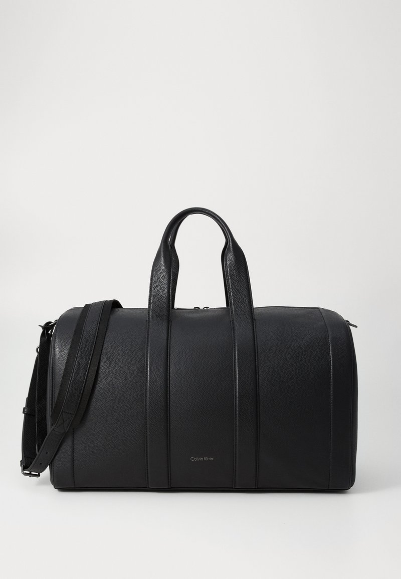 Bolsa de viaje de piel negra con asas dobles, correa para el hombro y rayas elevadas. Presenta el logo de Calvin Klein en el frente. Textura suave, forma redondeada.