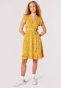 Gelbes Blumenkleid mit kurzen Rüschensäumen, einem V-Ausschnitt, tailliertem Bund, seitlichem Bindeband und einem gestuften Saum, kombiniert mit weißen Sneakers.