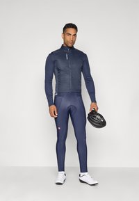 Chaqueta de ciclismo azul marino con cuello alto, mangas largas y diseño acolchado; combinada con leggings ajustados a juego y zapatillas de ciclismo blancas.