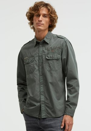 Junger Mann mit lockigem Haar, der ein graues Hemd mit Knöpfen, zwei Brusttaschen und gesticktem Schriftzug trägt und mit den Händen in den Taschen steht.