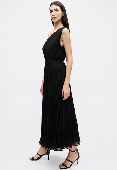 Γυναίκα που φοράει μαύρο πλισέ μάξι φόρεμα με ένα ώμο και μαύρα ψηλοτάκουνα σανδάλια με λουράκια, στέκεται μπροστά σε ένα απλό λευκό φόντο.