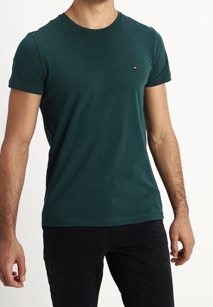 Mężczyzna w dopasowanym, ciemnozielonym t-shircie z krótkim rękawem i małym logo na klatce piersiowej oraz czarnych spodniach, stojący na jednolitym tle.