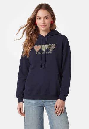 Ung kvinde med langt hår iført røde hjerteøreringe, en navy hoodie med tre stribede hjerter og tekst, samt lyseblå jeans mod en hvid baggrund.