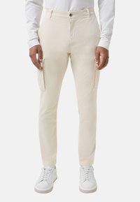 Mann, der off-white Slim-Fit Cargo-Hosen mit Seitentaschen und weißen Sneakers trägt, gezeigt von der Taille bis zu den Füßen vor einem einfarbigen Hintergrund.