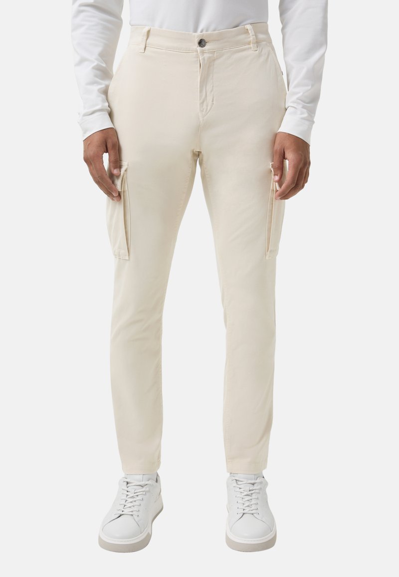 Mann, der off-white Slim-Fit Cargo-Hosen mit Seitentaschen und weißen Sneakers trägt, gezeigt von der Taille bis zu den Füßen vor einem einfarbigen Hintergrund.
