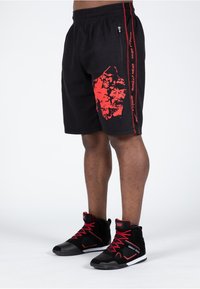 Schwarze Sportshorts mit strukturierter Oberfläche, die mit einem roten Grafikdruck und seitlichen Streifen versehen sind. Kombiniert mit schwarzen High-Top-Sneakers mit roten Akzenten.