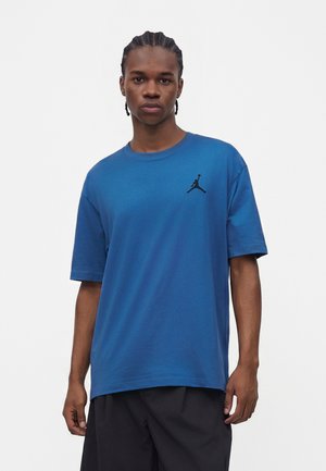 Młody mężczyzna z warkoczami, ubrany w niebieską koszulkę z krótkim rękawem z małym czarnym logo Jumpman na piersi.