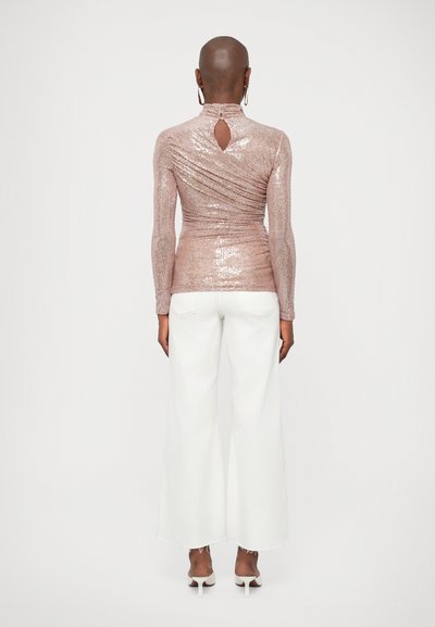 Μπλούζα με ψηλό λαιμό και μακριά μανίκια από λαμπερό ροζ χρυσό ύφασμα, με λεπτομέρεια κλειδώματος στην πλάτη, συνδυασμένη με φαρδιά λευκά παντελόνια και λευκές γόβες.