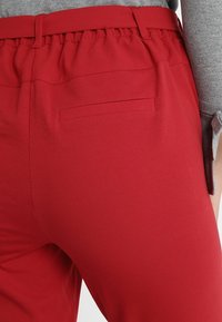 Røde bukser med elastisk talje. Har en baglomme med ren finish og glat tekstur. Viser også en ærme fra en grå trøje.