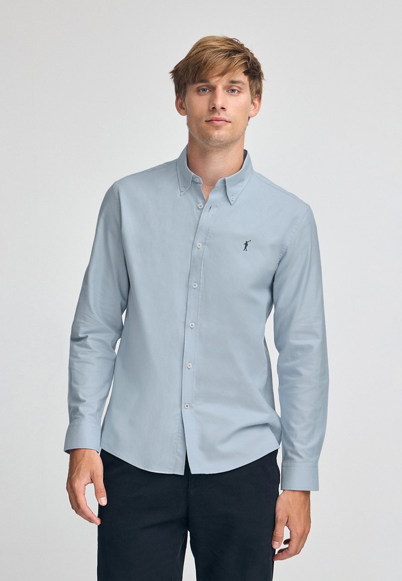 Ljusblå skjorta med knappar i bomull, med en liten mörk logotyp på bröstet, långa ärmar och standardkrage.