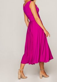 Robe sans manches fuchsia, longueur genou, en tissu lisse avec une jupe évasée. Associée à des sandales à talons carrés couleur nude.
