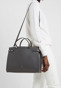 Henkilö, joka on pukeutunut valkoiseen neulepaitaan ja housuihin, kantaa suurta tummanharmaata nahkakäsilaukkua, jossa on olkahihna ja pienet kahvat.