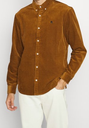 Hombre con camisa marrón de pana abotonada con botones blancos y pantalones de color claro, con la mano en el bolsillo.