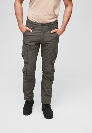 Mann trägt dunkelgraue Cargohosen und schwarze Stiefel, mit den Händen in den Taschen, steht vor einem schlichten grauen Hintergrund.