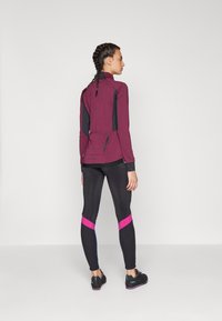 Burgundi sportdzseki fekete részletekkel, magas gallérral és hátoldali cipzárral. Fekete leggings rózsaszín és kék ferdén futó csíkokkal. Fekete lábbelik.
