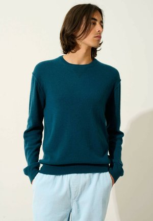 Türkiser Pullover mit langen Ärmeln, rundem Ausschnitt und strukturiertem Detail am Kragen, kombiniert mit hellblauen gestreiften Hosen.