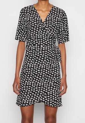 Woman wearing a black short-sleeve wrap dress with a white floral pattern, tied at the waist, standing against a plain background.