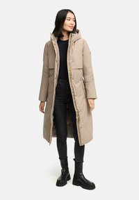 Beige lang pufferjakke med hætte, trykknapper, sidelommer og quiltet inderside, parret med sorte jeans og støvler.