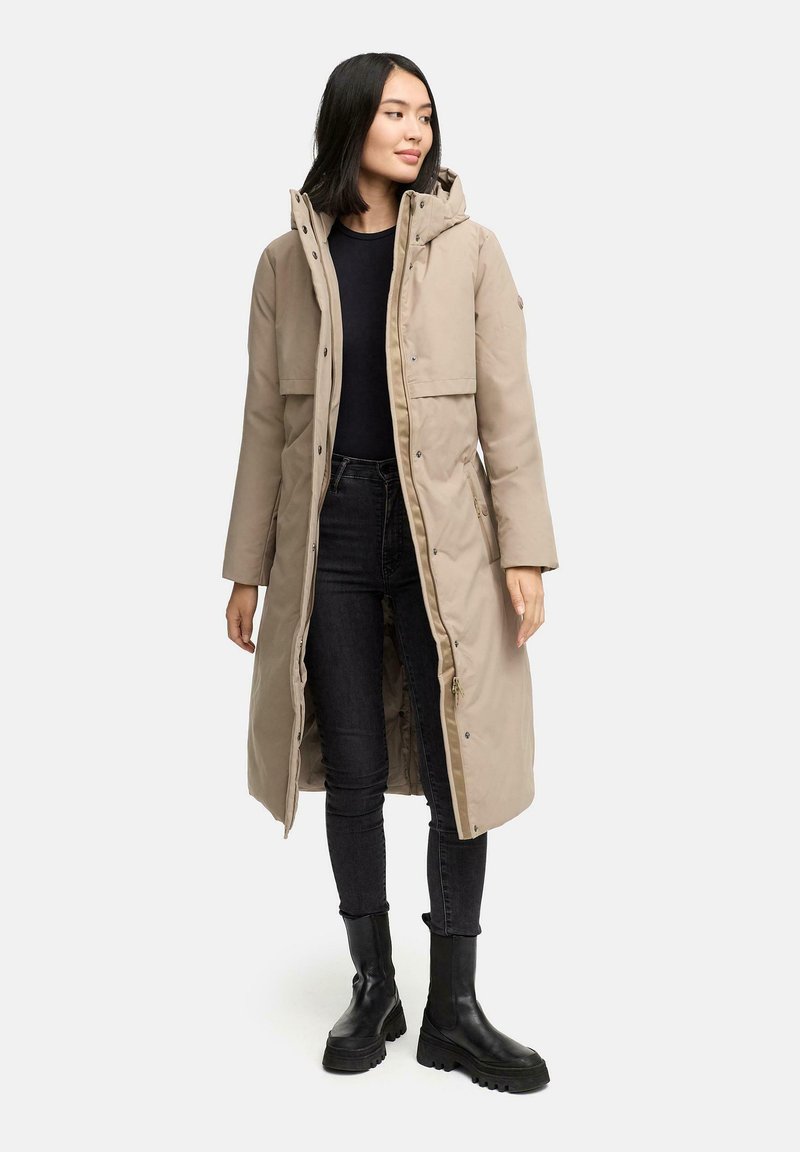 Beige lang pufferjakke med hætte, trykknapper, sidelommer og quiltet inderside, parret med sorte jeans og støvler.