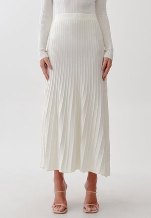 Femme portant un haut blanc côtelé à manches longues, une jupe midi plissée blanche et des sandales à talons hauts nude à brides, debout devant un fond uni.