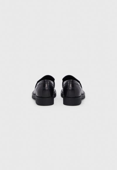 Ζευγάρι μαύρα δερμάτινα παπούτσια επίσημου τύπου, ορατά από το πίσω μέρος, με χαμηλά τακούνια και εμφανείς ραφές σε απλό φωτεινό φόντο.