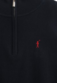Marinblå stickad halvzip-tröja med en röd broderad figurlogo på vänster bröst, med en hög ribbad krage och slät textur.