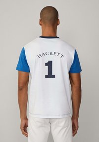 Bílý a modrý krátký rukáv tričko s nápisem "HACKETT" a číslem "1" na zádech, se kulatým výstřihem a prošitým lemem.