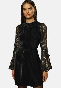 Rochie neagră din catifea, cu mâneci din tul brodate cu flori și volane la manșete, având un gât strâns și talie accentuată.
