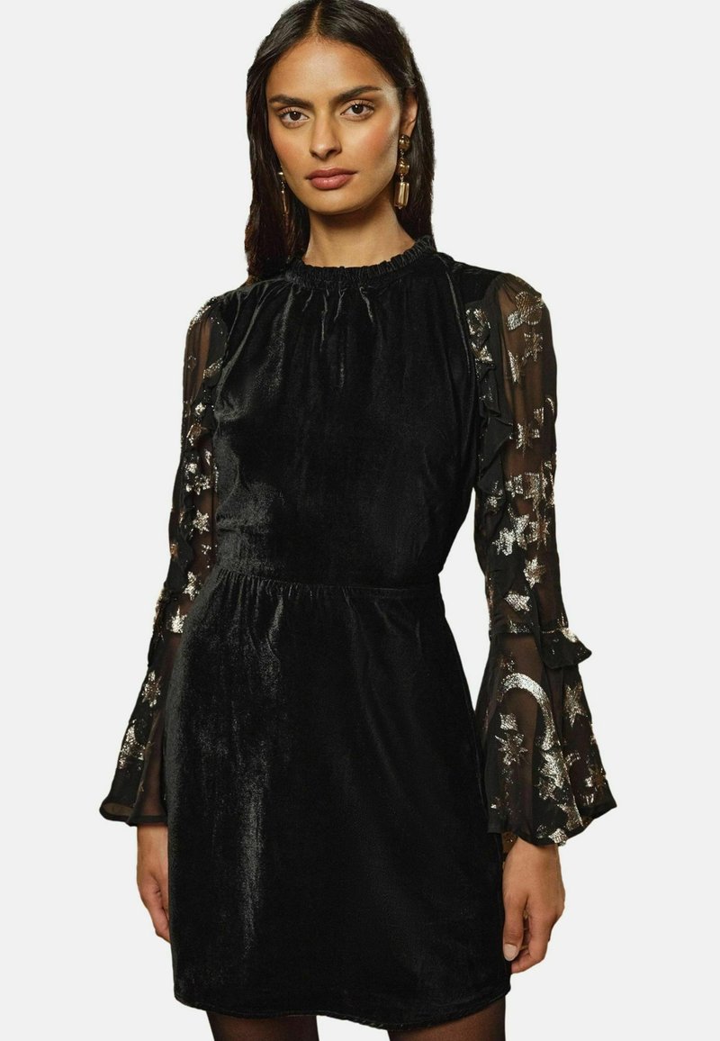 Rochie neagră din catifea, cu mâneci din tul brodate cu flori și volane la manșete, având un gât strâns și talie accentuată.