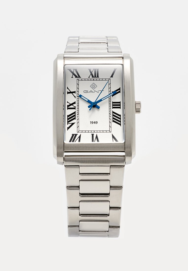 Montre rectangulaire argentée avec un cadran blanc, des chiffres romains noirs et des aiguilles bleues. Équipée d'un bracelet en maillons métalliques et du logo GANT.