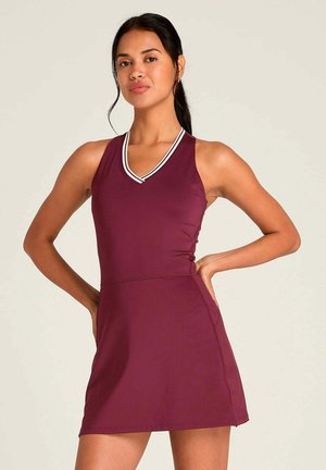 Frau mit dunklem Haar in einem Pferdeschwanz, die ein ärmelloses dunkelrotes Kleid mit weiß gestreiftem V-Ausschnitt trägt und mit den Händen in die Hüften gestützt steht.