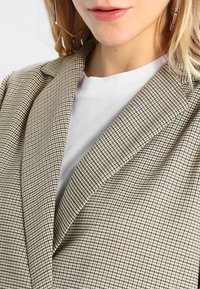 Marynarka w kratę typu pepitka w beżowo-czarnym materiale, z wyłogami szpiczastymi. Nałożona na gładki biały t-shirt.