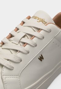 Λευκό δερμάτινο αθλητικό παπούτσι με λευκά κορδόνια, μπεζ λεπτομέρειες, χρυσό λογότυπο "Wrangler" στη γλώσσα και μικρό χρυσό "W" στο πλάι.